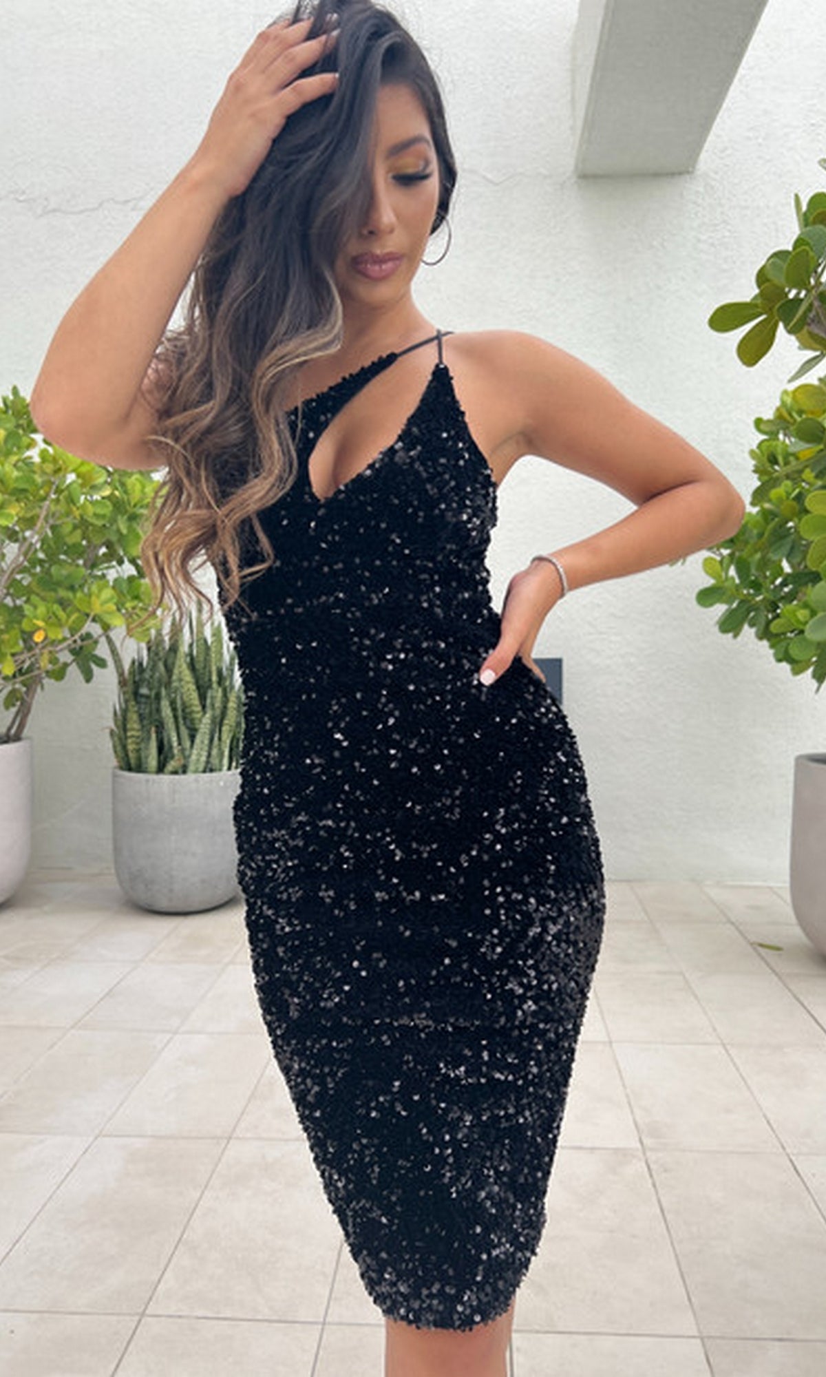 A woman poses in the Venice One-Shoulder Short Black Sequin Party Dress on a tiled floor, touching her hair, with potted plants in the background.